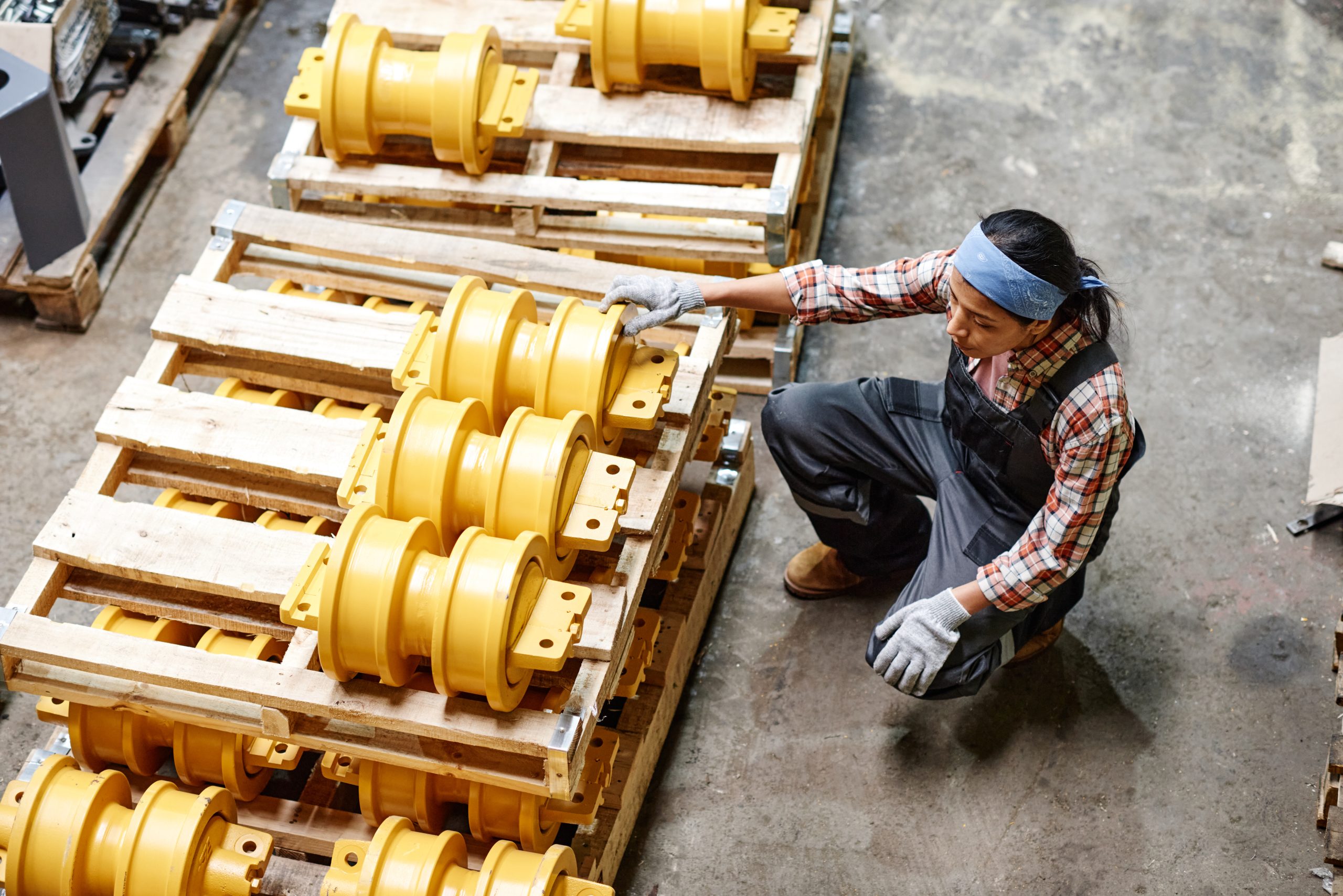 Above angle of young female worker of industrial plant sitting by stack of spare parts while choosing working material for machine repair