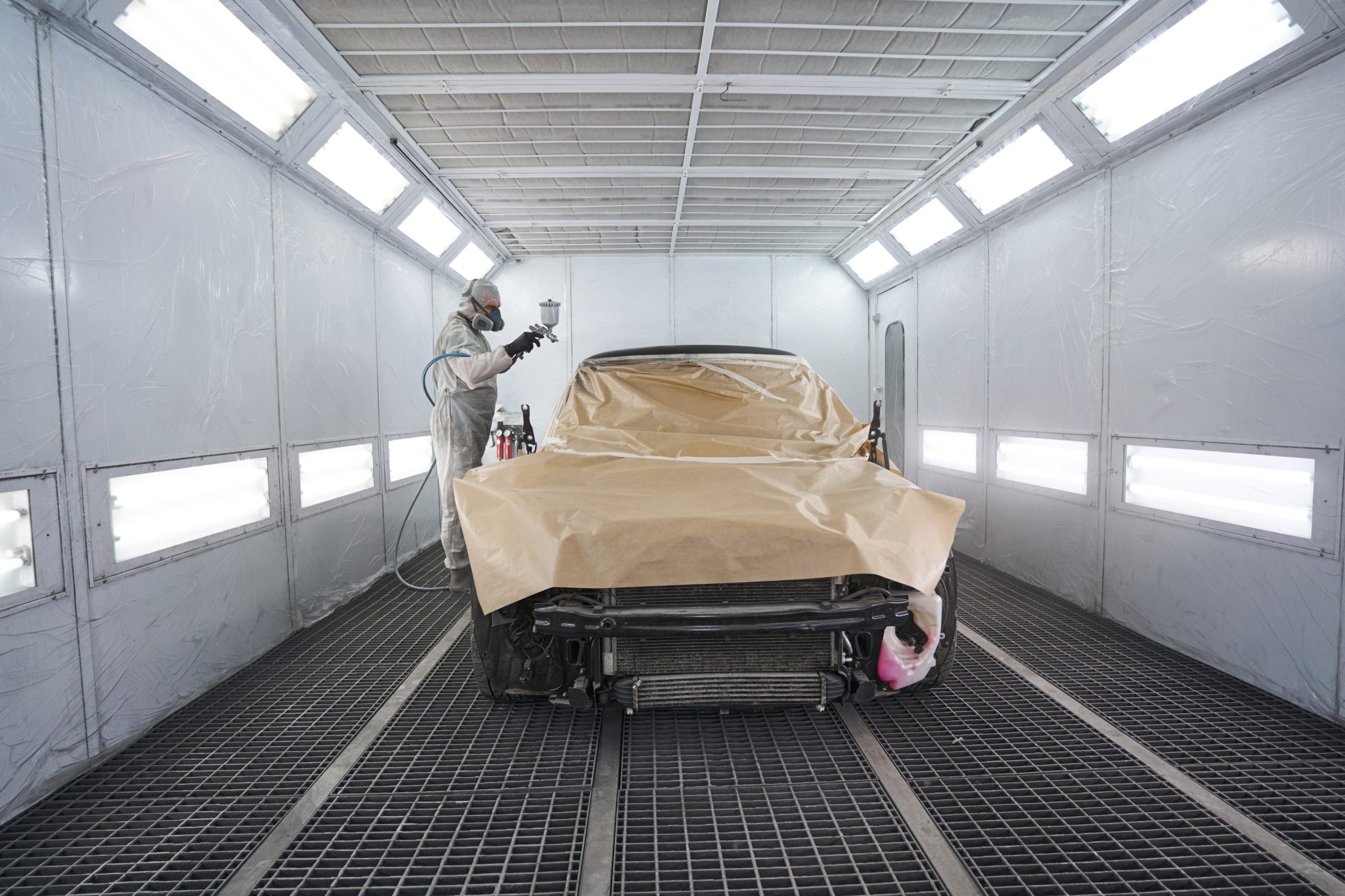 Car painter in white protective clothes, gloves and respirator standing in car painting booth, holding spray gun and working
